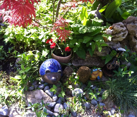 A vibrant garden scene features a mix of lush green foliage and bright red flowers. Among the plants are decorative garden ornaments, including a blue hedgehog figurine and a whimsical frog figure. The setting includes natural rocks and pebbles, adding texture to the scene.